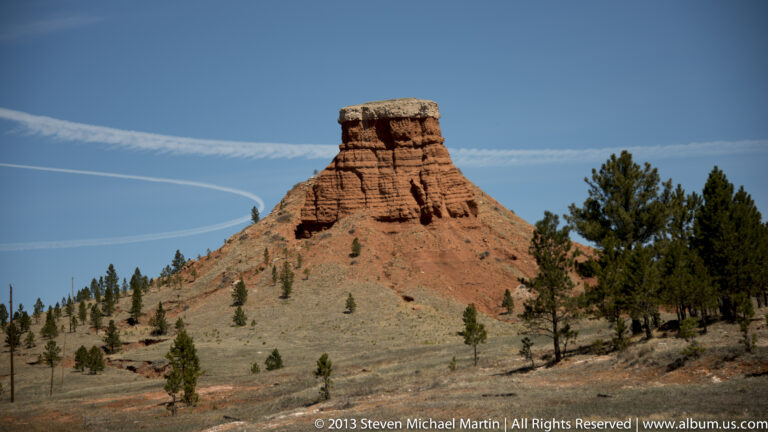 A red, banded butte with a flat pale cap rises above a grassy slope dotted with pine trees.