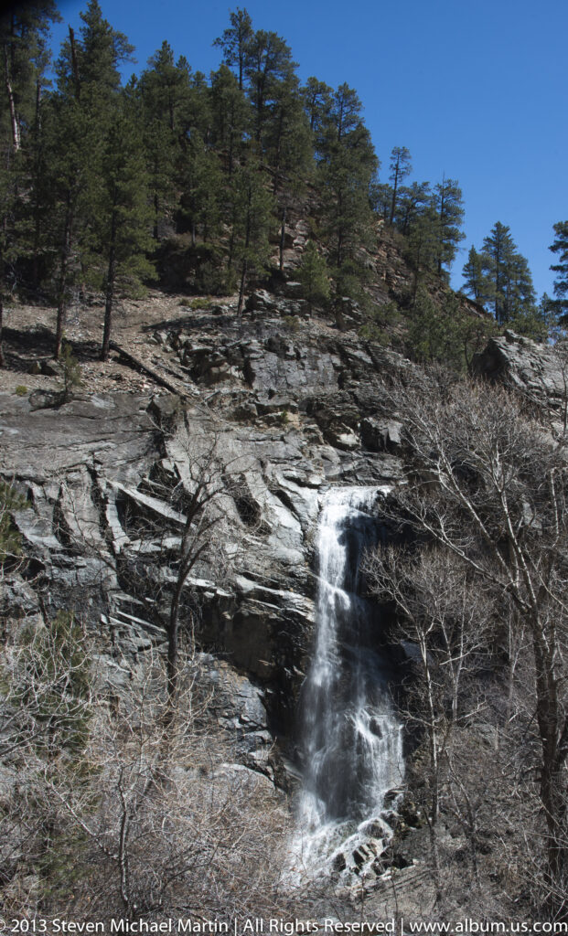 A small waterfall spills over layered dark rock beneath pines and leafless shrubs.