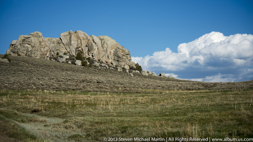 A low granite ridge rises above open grassland beneath blue sky and white clouds.