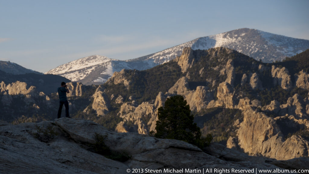 A visitor on a granite overlook looks across rocky ridges toward snow-dusted peaks.