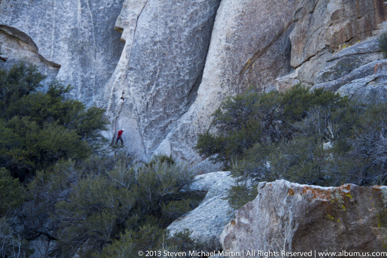 A climber in a red top ascends a broad granite slab above shrubs.
