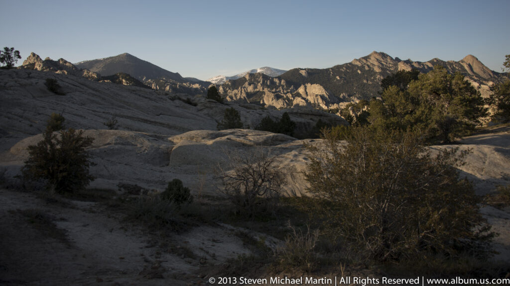 A broad granite shelf and scattered shrubs lead toward distant rugged peaks.