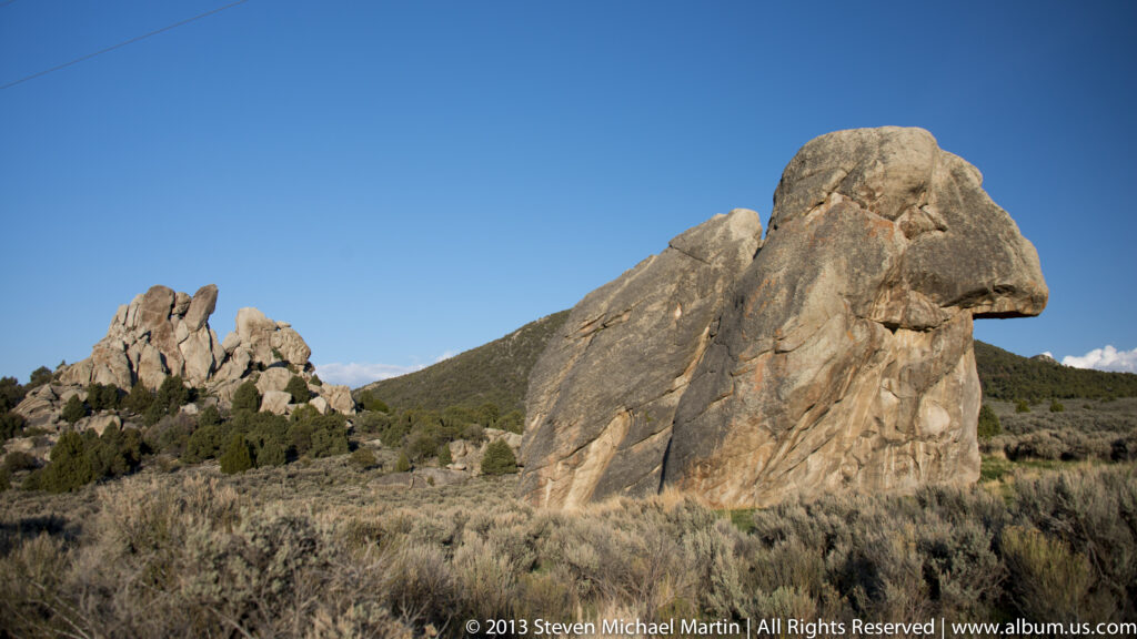A cap-shaped granite boulder stands before distant spires and low sagebrush.