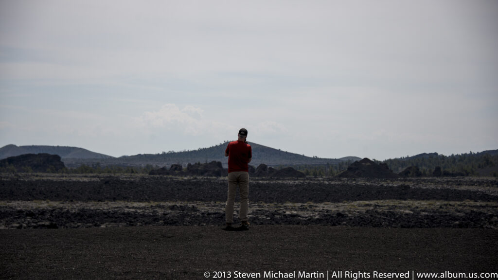 A visitor in red faces a broad dark lava field, with low ridges on the horizon.