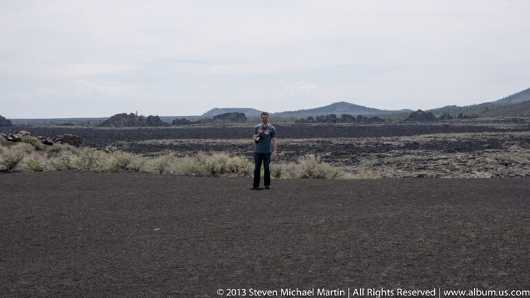 A lone visitor stands on dark volcanic gravel before a lava field and distant hills.