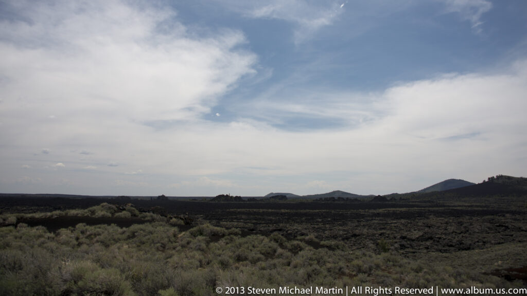 Sagebrush and grasses border a broad dark lava field beneath a cloud-streaked sky.