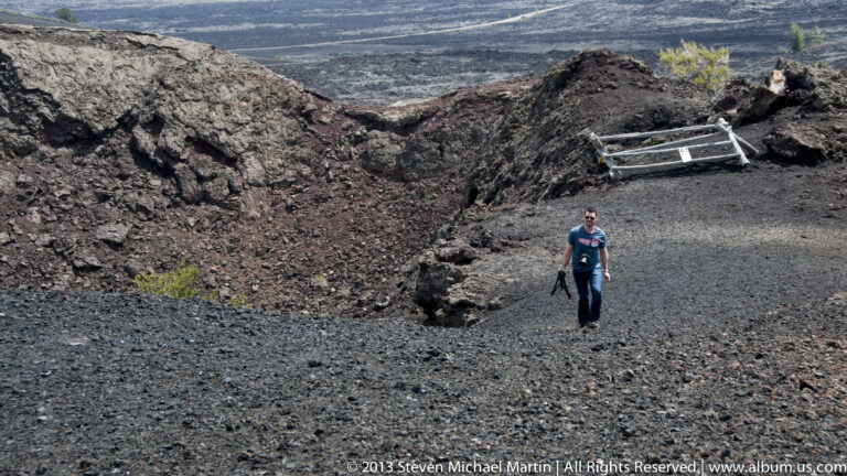 A visitor walks across dark cinders below a reddish crater wall and metal platform.