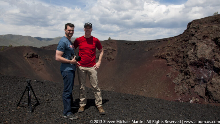 Two visitors stand on dark cinders inside a red-brown volcanic crater.