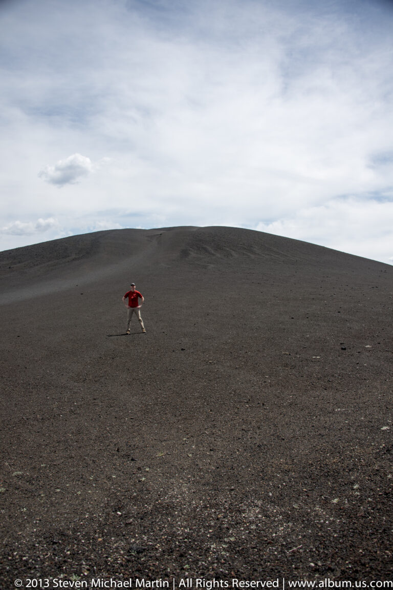 A small visitor crosses a broad dark cinder slope beneath a cloudy sky.