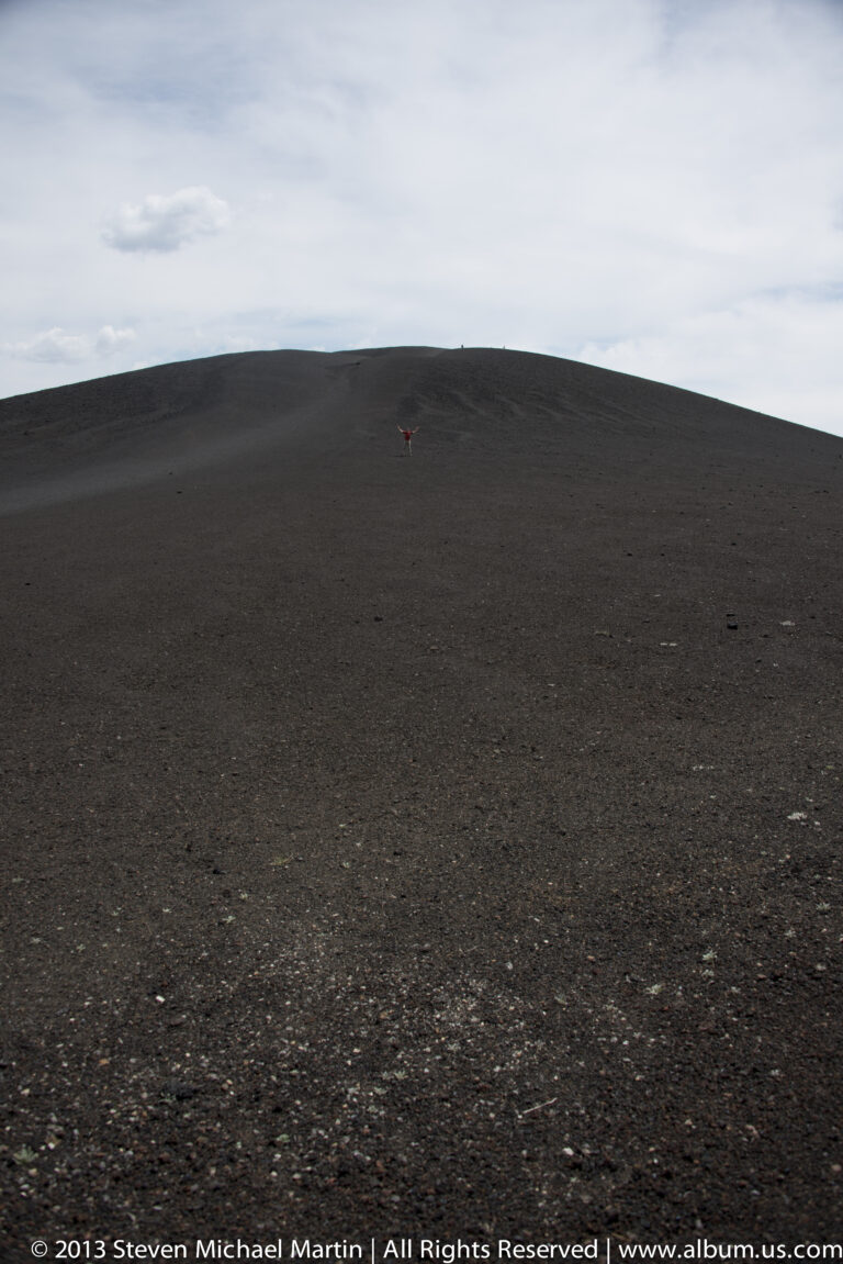 A tiny red figure stands near the crest of a broad dark volcanic hill.