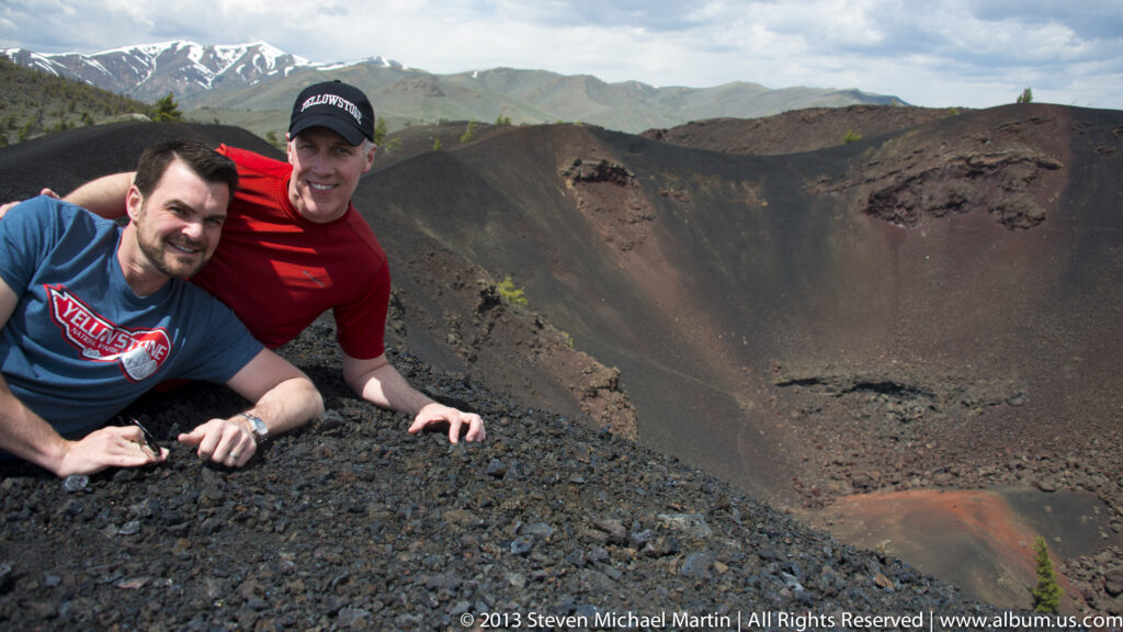 Two visitors lean over a black cinder rim above a broad red-brown crater.
