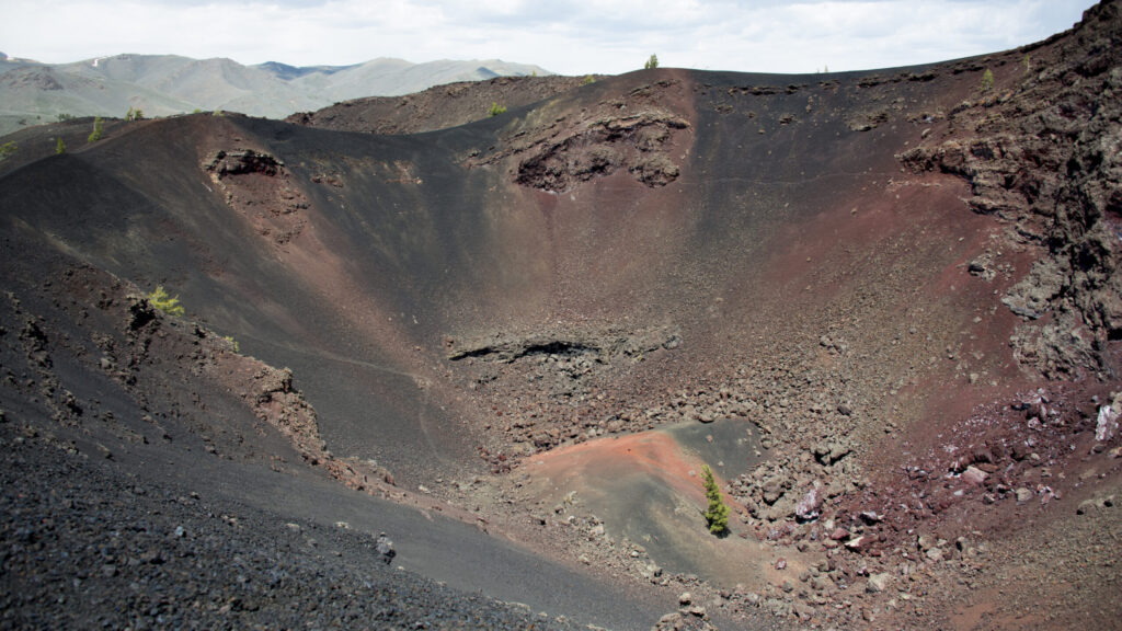 A wide volcanic bowl has a dark rim, reddish inner slopes, and sparse plants.