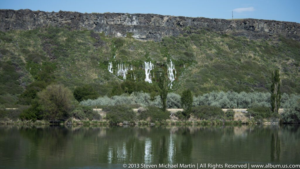Multiple narrow waterfalls descend a green canyon wall above a calm reflective river.