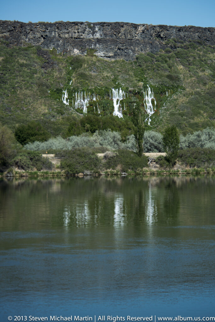 White waterfalls cross a broad green canyon slope above a blue river.