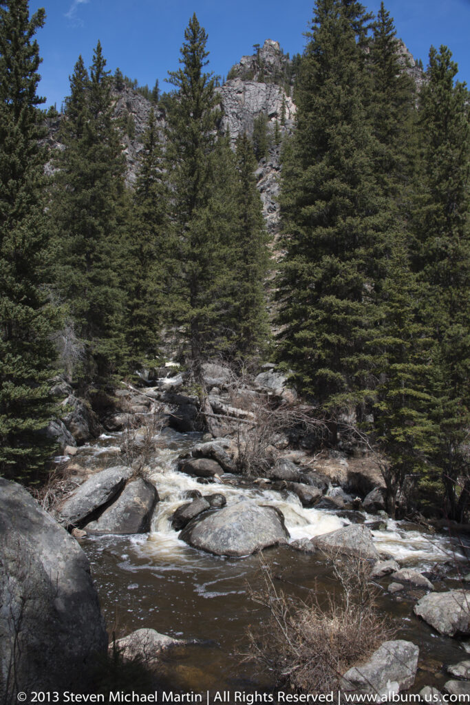 A rocky stream flows between conifer-covered slopes beneath an exposed cliff.