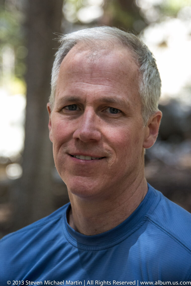 A gray-haired man in a blue shirt looks toward the camera against a softly blurred wooded background.