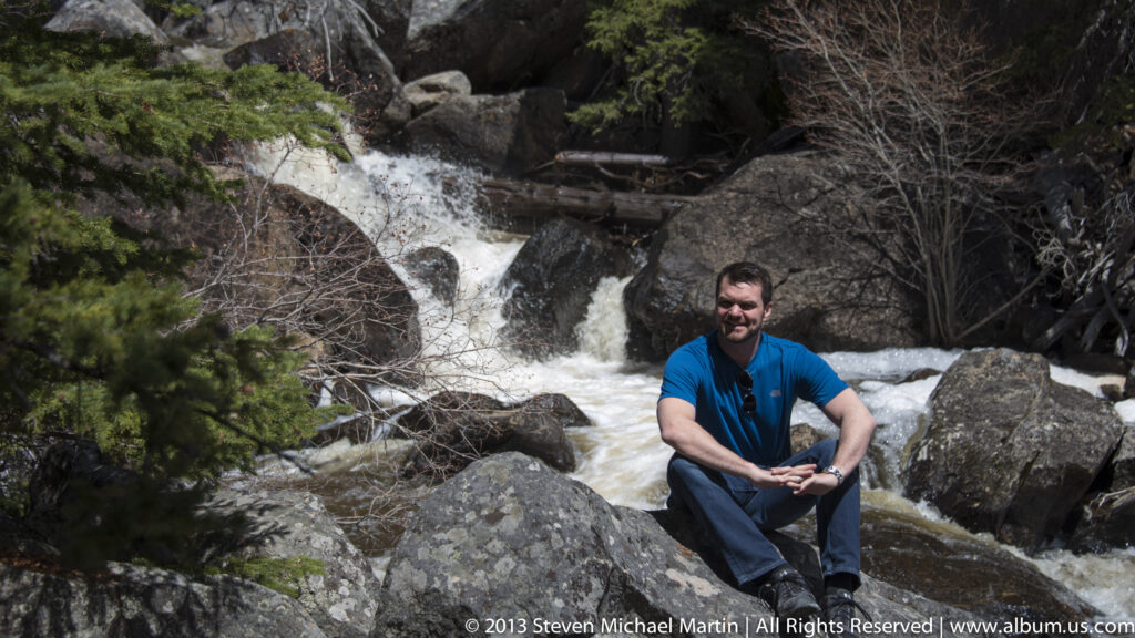 A visitor sits on a boulder beside a rushing cascade framed by trees.