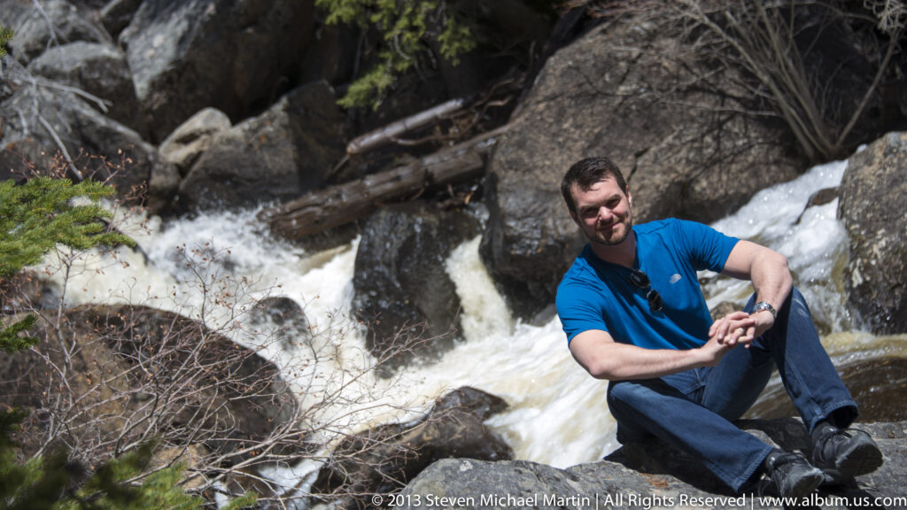 A seated visitor rests beside foaming water rushing over boulders in a wooded channel.