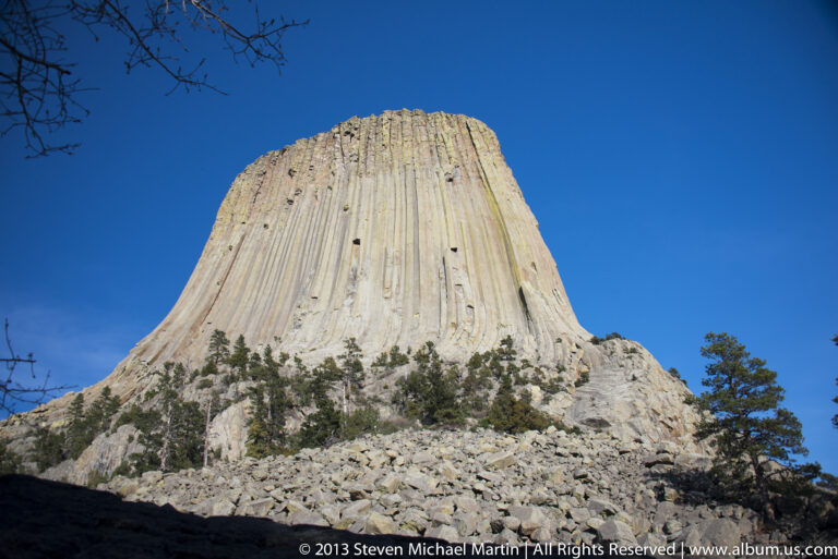 Devils Tower rises above a boulder-covered slope, its vertical columns set against a blue sky.