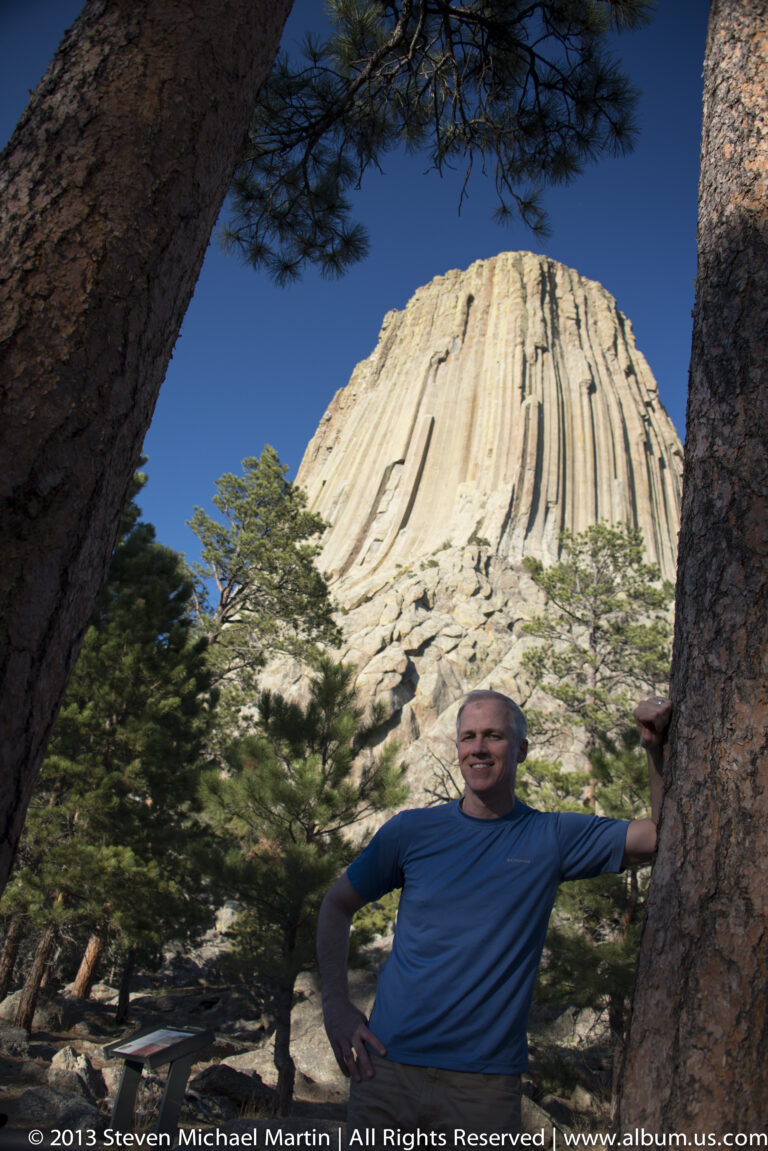 A visitor stands beside a pine beneath Devils Tower, with the formation towering behind him.
