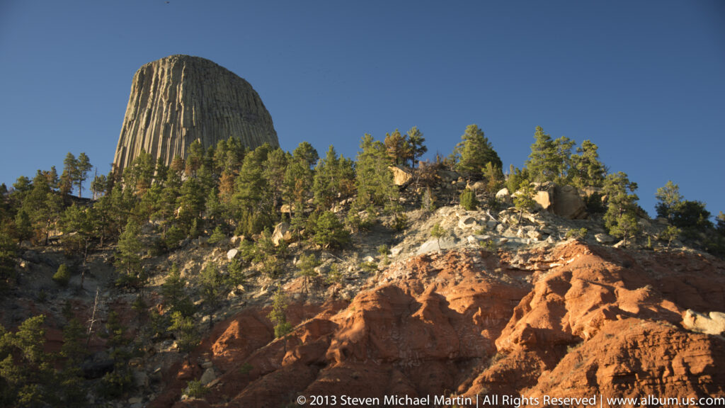 The upper columns of Devils Tower rise above pines and a reddish rocky slope.