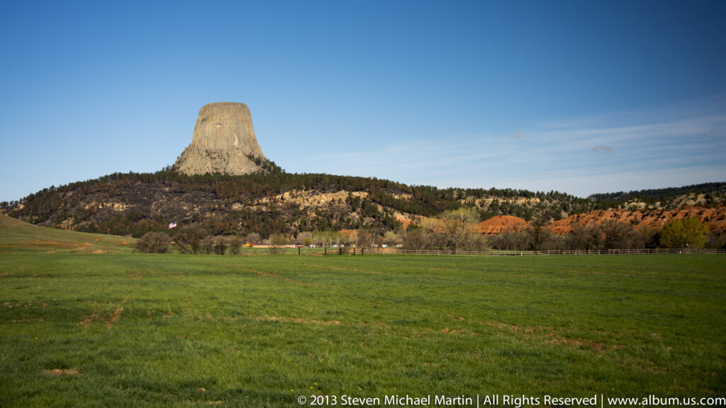 Devils Tower rises beyond a green meadow and autumn-colored trees.