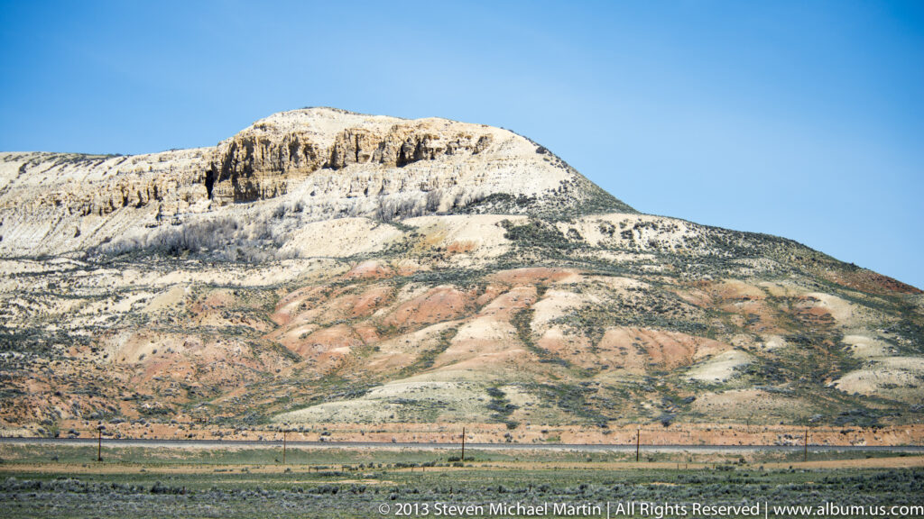 A pale layered cliff rises above rust-colored slopes and sparse vegetation beneath a clear sky.