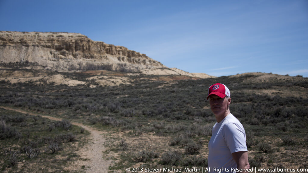 A visitor stands beside a trail below pale layered cliffs and low shrubs.