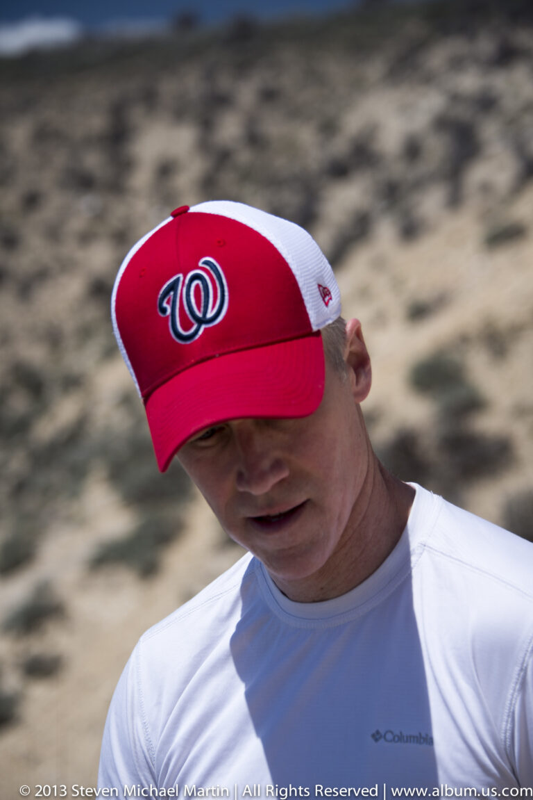 A man in a red Washington cap and white shirt looks downward against a blurred rocky background.