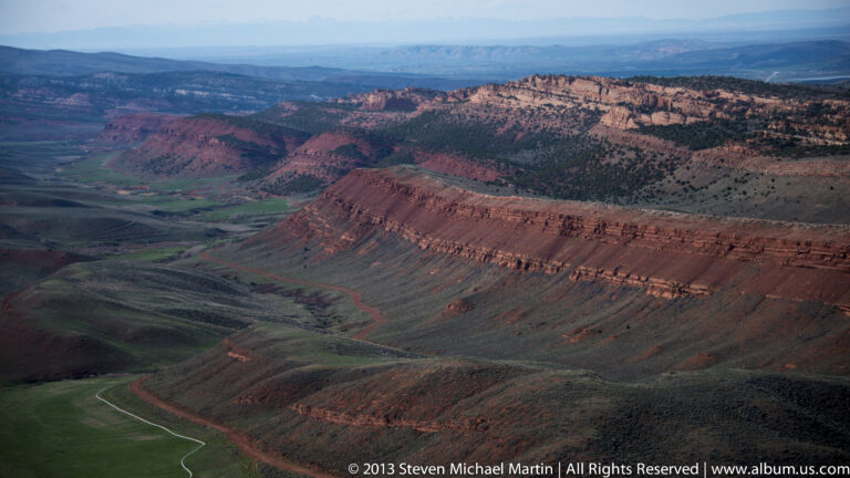 An elevated view shows red ridges, a green basin, and a pale road winding toward distant hills.