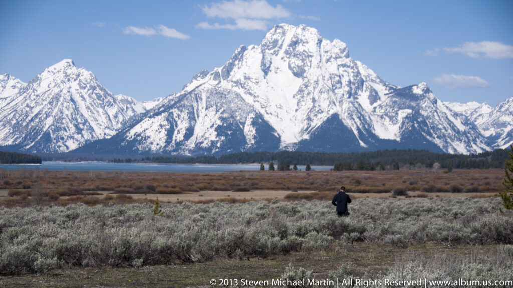 A lone person stands in sagebrush below snow-covered peaks across a strip of water.