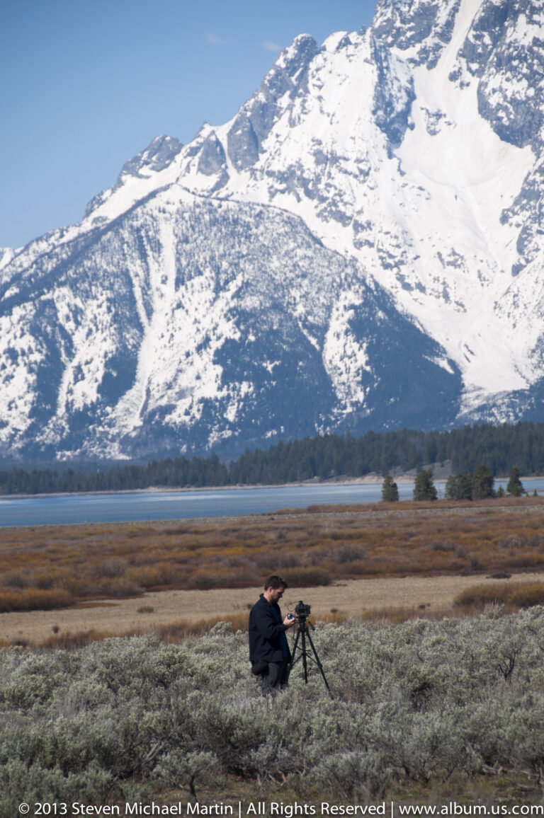 A photographer crouches at a tripod in sagebrush with snowy mountains across the valley.