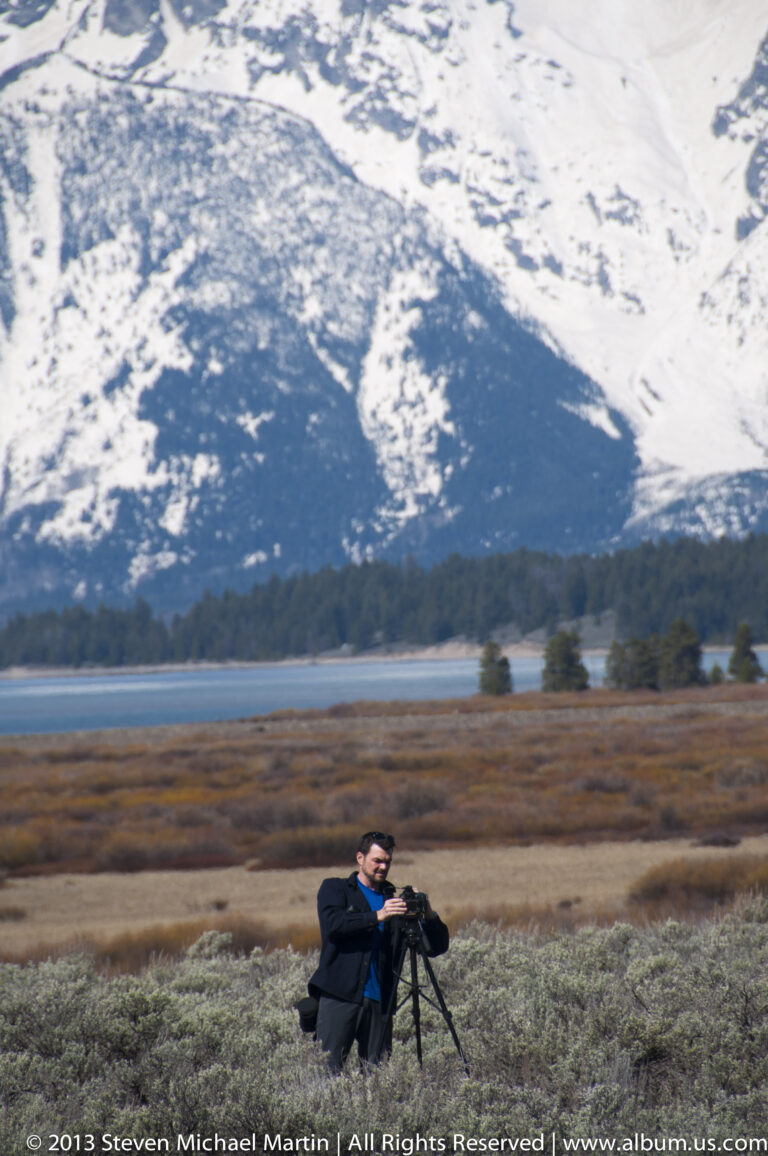 A seated photographer rests beside a tripod in sagebrush, with snowy mountains behind.