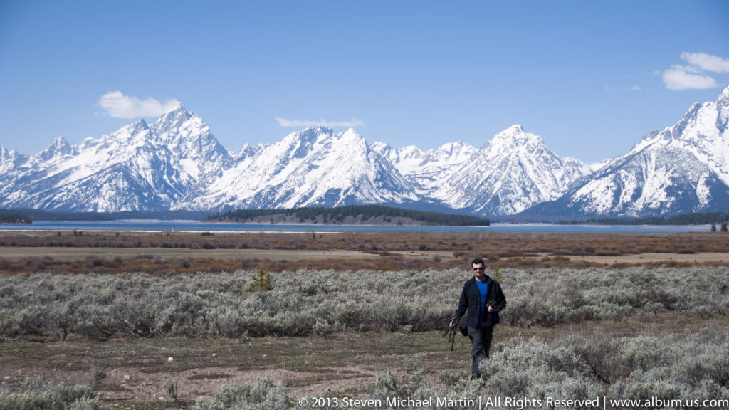 A man walks through sagebrush with a lake and snowy Teton peaks behind him.