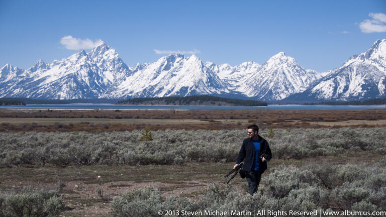 A man crosses scrubby sagebrush with the Teton Range stretching across the background.
