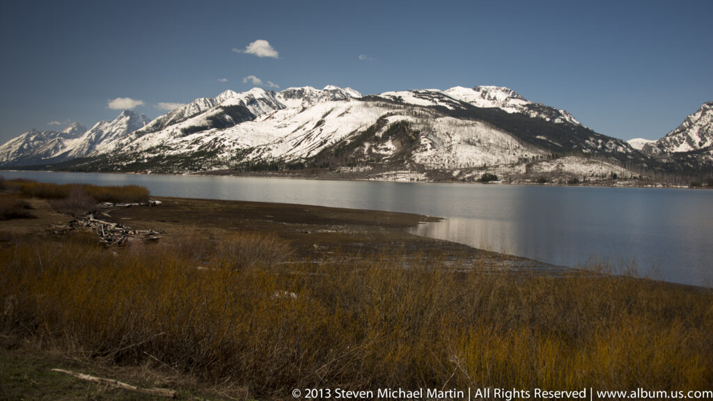 A lake lies below a snow-dusted ridge, beyond tawny marsh grasses and low shrubs.