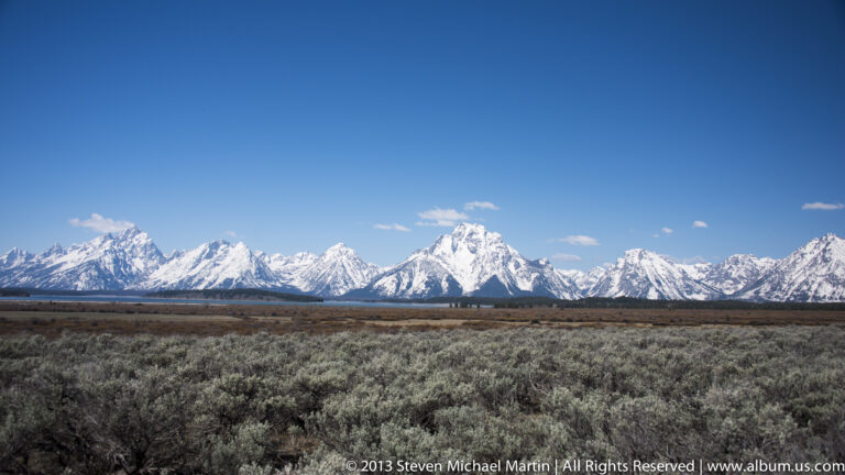 A broad sagebrush plain stretches toward the snow-covered Teton Range under a blue sky.