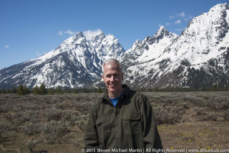 A man stands in sagebrush with the snow-covered Teton Range behind him.