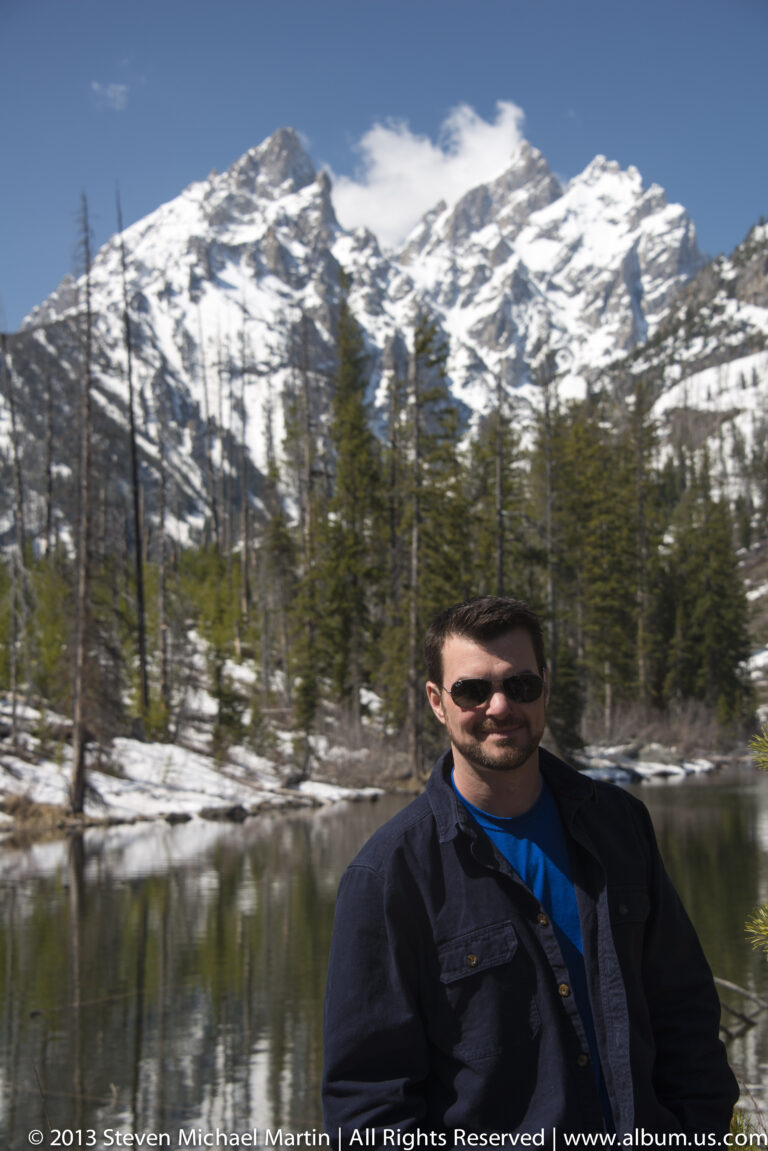 A seated man poses beside a pond, with conifers and patches of snow behind him.