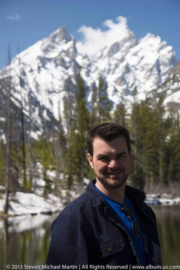 A close portrait of a man beside a pond with snow and evergreens behind him.