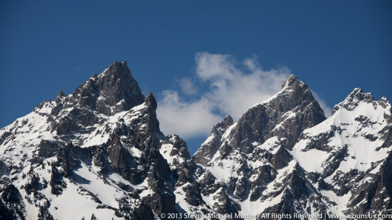Jagged dark peaks with snow-filled gullies rise beneath a blue sky and small cloud.