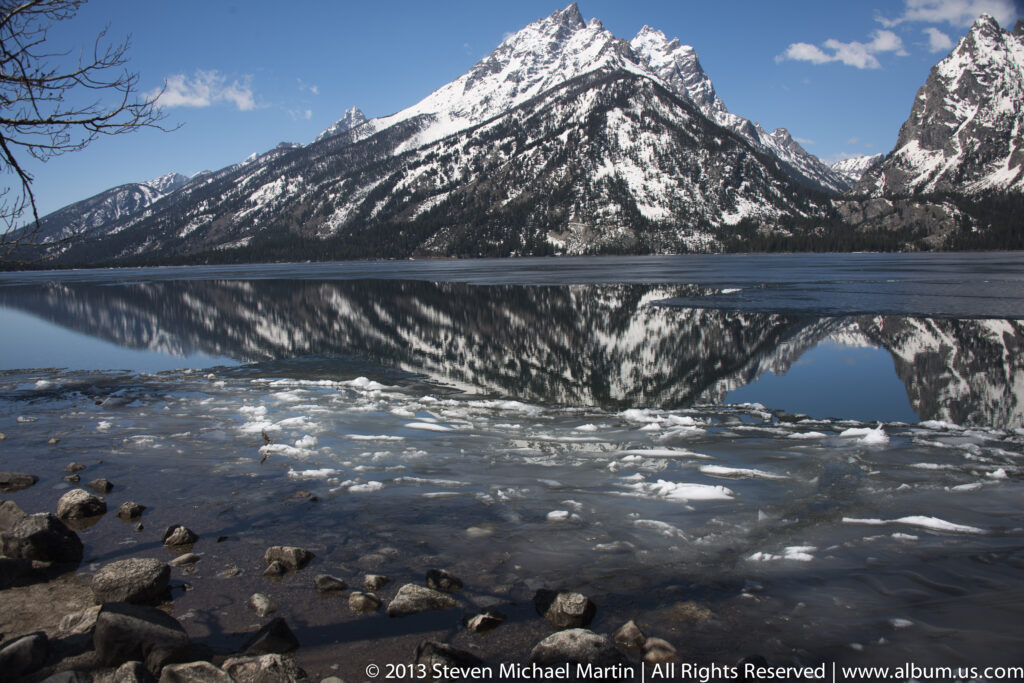 Ice floes and shoreline rocks sit before a lake reflecting a snowy mountain.