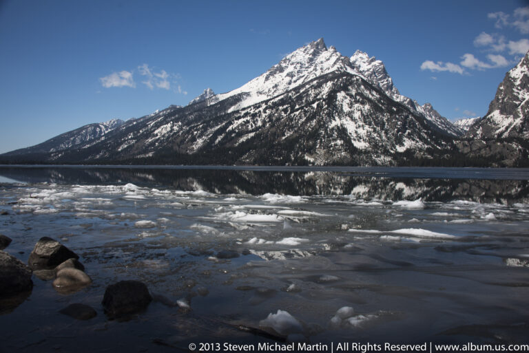 A snow-covered peak and forest reflect in a lake dotted with small pieces of ice.