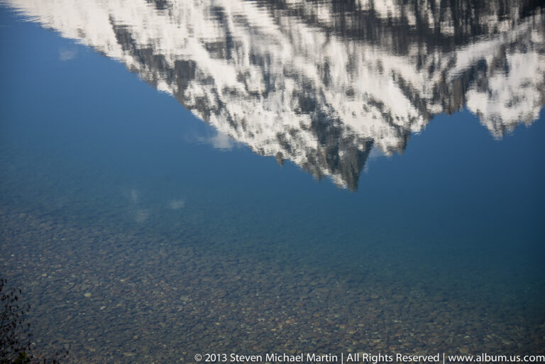 Snow-covered peaks appear as a reflection in deep blue lake water, seen from close to shore.