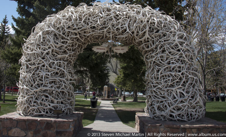 An arch of pale antlers spans a park path between stone supports.