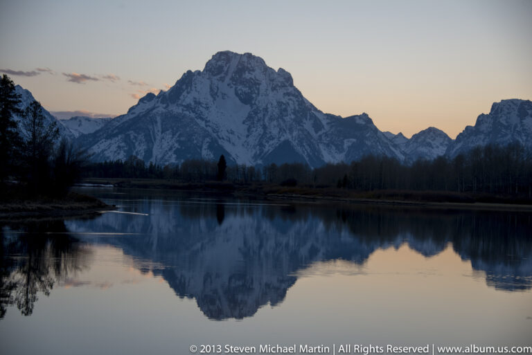 A snow-lined mountain and pastel dawn sky reflect in a still lake.