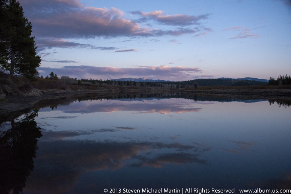 Pink and lavender clouds reflect in a still wetland bordered by dark trees and grasses.
