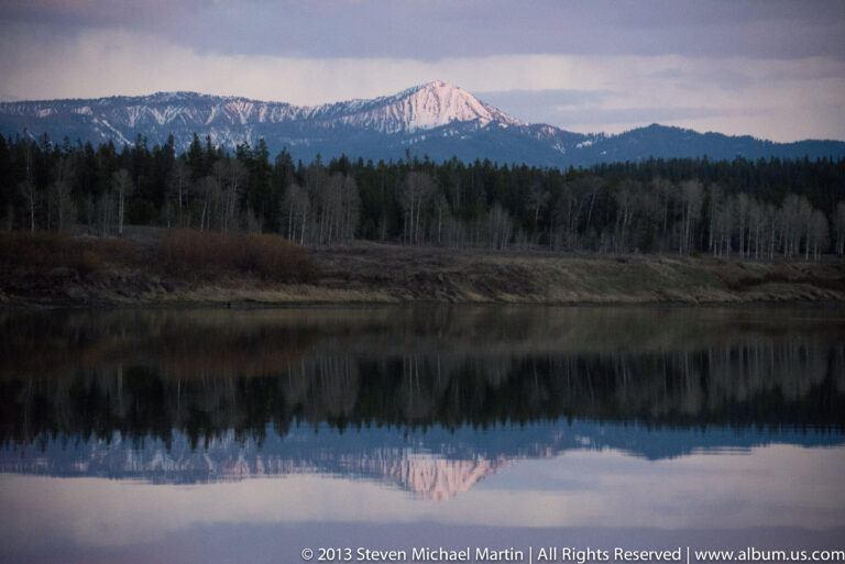 A pink-lit, snow-streaked Teton peak and forested shoreline reflect in a still lake.