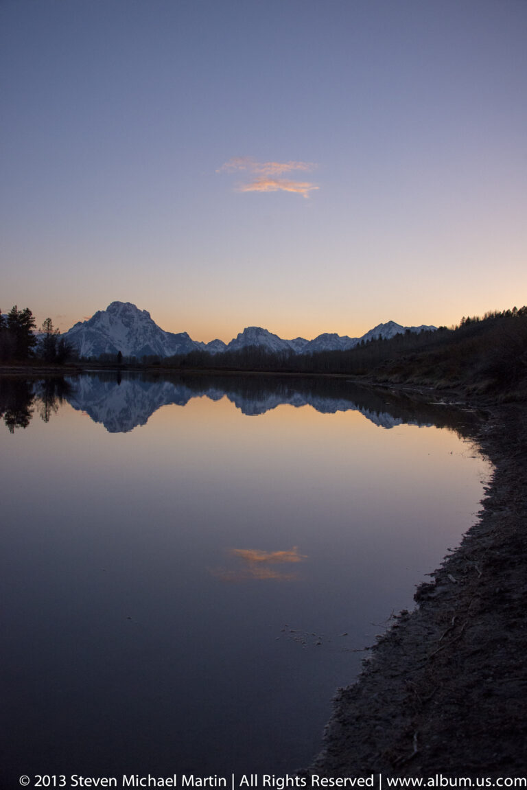 A low snowy ridgeline and pastel sunset sky reflect across a still lake beside a dark shore.