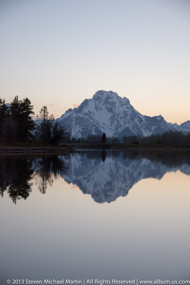 A broad snowy mountain and dark treeline are reflected in a calm lake under a pale evening sky.